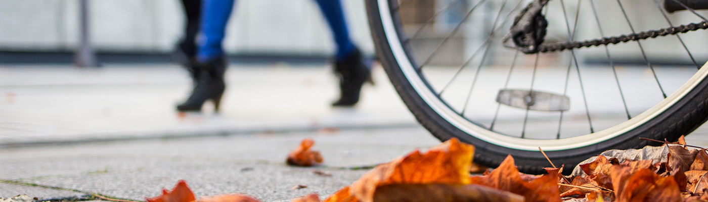 A bike wheel, a woman walking and some leaves on the ground
