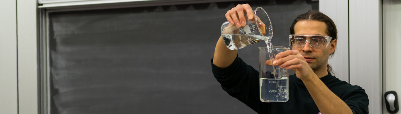 Lecturer pouring chemicals between beakers in front of a blackboard