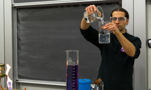 Lecturer pouring chemicals between beakers in front of a blackboard