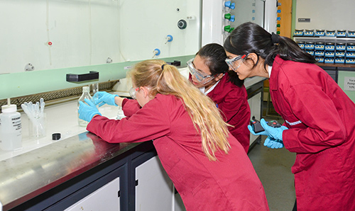 Three school children in red coats observing experiment in lab