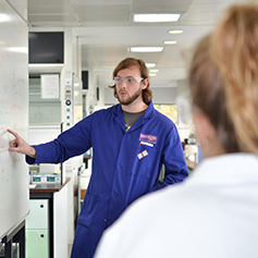 A male researcher in blue lab coat pointing to a whiteboard
