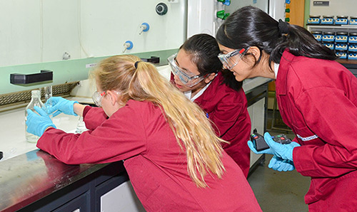 Three school children in red lab coats observe experiment in lab