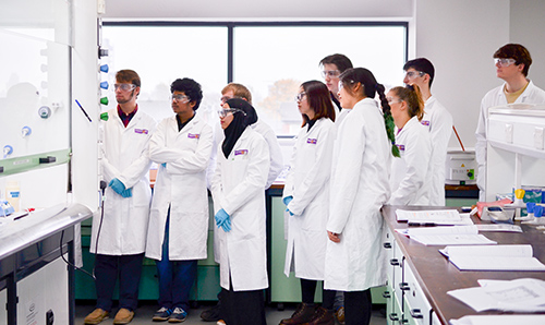 A row of students wearing white lab coats and goggles observe an experiment in a lab