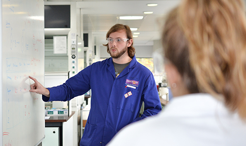 Student in blue lab coat explains content of whiteboard