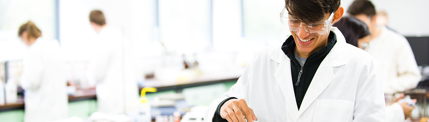 A male student in a lab coat smiling as he works on an experiment